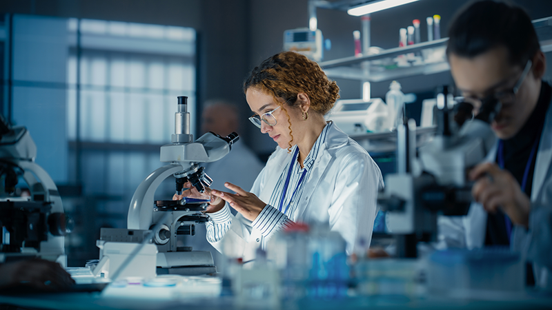 A female bioengineer examining DNA samples under a microscope in a modern applied science laboratory. 